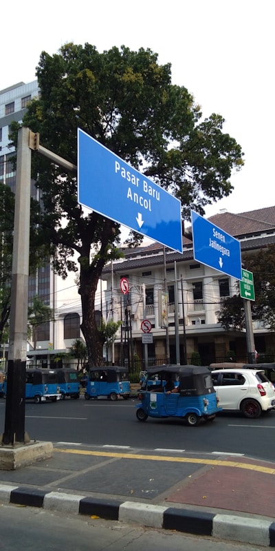 Several blue autorickshaws are parked along a street with large blue road signs pointing toward Pasar Baru and Ancol. The background features trees and a multi-story building.