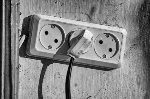 A black and white photo of a double electrical outlet on a worn, textured wall. One of the sockets is occupied by a plug with a visible cord hanging down. The surrounding area is slightly shadowed, emphasizing the plug's prominence.