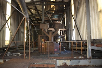 An industrial interior with metal beams, pipes, and machinery. The environment appears aged and rusted, with panel walls and a grated metal floor. Light filters through narrow windows, casting shadows.