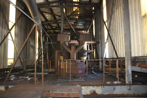 An industrial interior with metal beams, pipes, and machinery. The environment appears aged and rusted, with panel walls and a grated metal floor. Light filters through narrow windows, casting shadows.