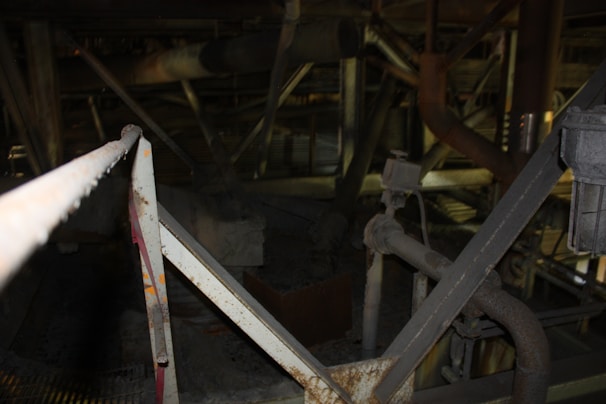 A dark and industrial environment with various metal beams, pipes, and machinery. The structures appear to be covered in a layer of dust or rust, indicating an aged and possibly abandoned setting. The lighting is dim, with some light illuminating specific parts of the scene.