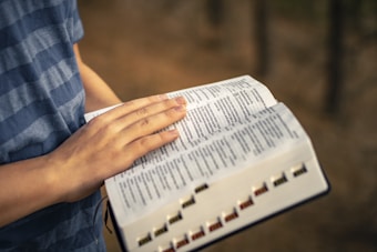 A person wearing a blue striped shirt is holding an open book and appears to be reading. The book's pages are open and visible, giving the impression of being studied or analyzed. The background is blurred, emphasizing the focus on the book and the reader's hands.