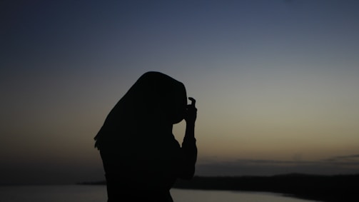 A silhouette of a person wearing a headscarf and holding a camera against a gradient sky during sunset or sunrise. The landscape includes a body of water and a distant horizon.