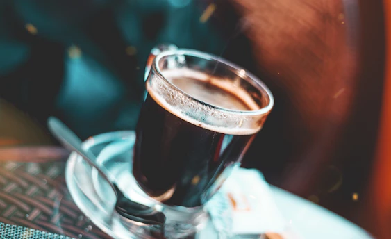 Close-up of a steaming cup of black coffee on a white table with a blurred Brooklyn street scene in the background.