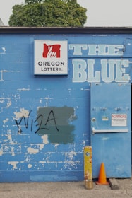 A weathered blue brick wall featuring a 'Oregon Lottery' sign with a stylized hand logo. The word 'BLUE' is painted in large letters next to a utility door with a 'No Minors Permitted' sign. There is graffiti reading 'VISA' on the wall, partially covering a green patch. An old yellow metal post and an orange traffic cone are positioned in front of the wall. A tree can be seen in the background above the wall.