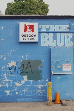 A weathered blue brick wall featuring a 'Oregon Lottery' sign with a stylized hand logo. The word 'BLUE' is painted in large letters next to a utility door with a 'No Minors Permitted' sign. There is graffiti reading 'VISA' on the wall, partially covering a green patch. An old yellow metal post and an orange traffic cone are positioned in front of the wall. A tree can be seen in the background above the wall.