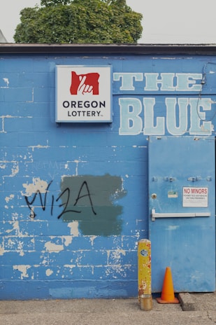 A weathered blue brick wall featuring a 'Oregon Lottery' sign with a stylized hand logo. The word 'BLUE' is painted in large letters next to a utility door with a 'No Minors Permitted' sign. There is graffiti reading 'VISA' on the wall, partially covering a green patch. An old yellow metal post and an orange traffic cone are positioned in front of the wall. A tree can be seen in the background above the wall.
