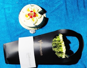 A round cake decorated with swirls of cream and red and yellow fruit sits on a bright blue surface. Beside it, a black packaging from a floral shop contains white flowers with green foliage, partially visible. There is also a small white card attached to the packaging.