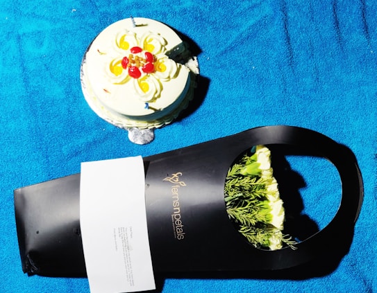 A round cake decorated with swirls of cream and red and yellow fruit sits on a bright blue surface. Beside it, a black packaging from a floral shop contains white flowers with green foliage, partially visible. There is also a small white card attached to the packaging.