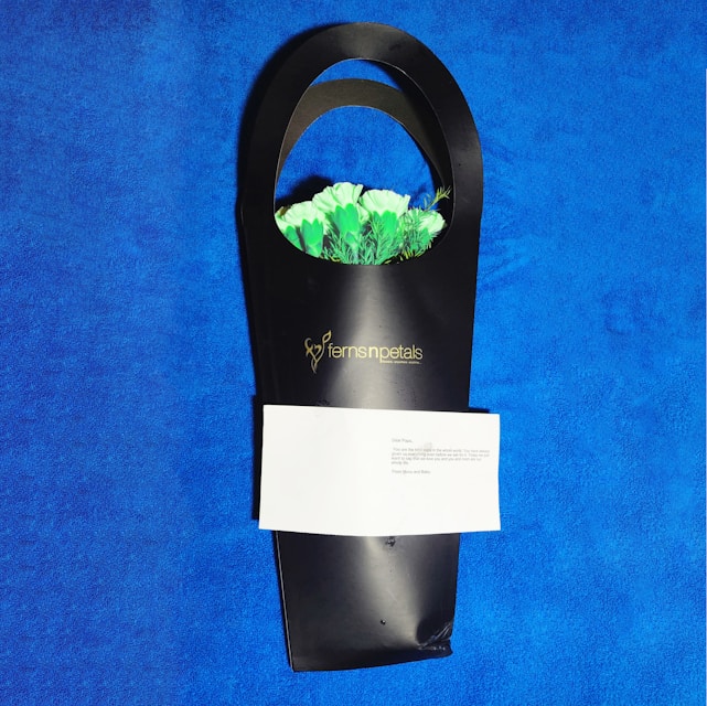 A sleek black gift bag holds a bouquet of white and green flowers with a visible logo reading 'ferns n petals'. There is a folded white note attached to the bag. The background is a bright, textured blue surface.
