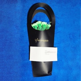 A sleek black gift bag holds a bouquet of white and green flowers with a visible logo reading 'ferns n petals'. There is a folded white note attached to the bag. The background is a bright, textured blue surface.
