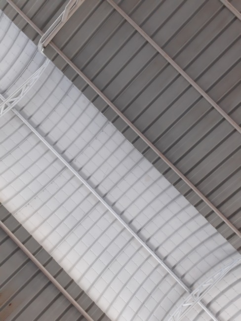 A ceiling with a prominent central duct running through it. The duct has a curved, grid-like pattern and is painted white. Surrounding the duct are gray metallic panels arranged in a linear fashion. The structure appears industrial with visible beams and metallic elements.