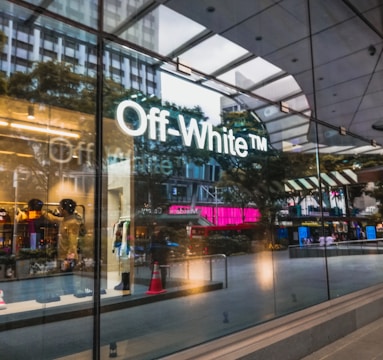 A storefront with large glass windows displaying the logo of Off-White. Inside, mannequins are arranged with clothing items. The reflection of nearby buildings and trees is visible on the glass. The atmosphere seems urban and modern, with a hint of outdoor activity visible through the glass.