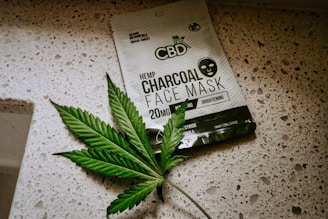A package of CBD hemp charcoal face mask lies on a speckled countertop surface. Next to the package is a green leaf with a serrated edge, resembling a cannabis leaf.