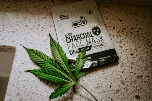 A package of CBD hemp charcoal face mask lies on a speckled countertop surface. Next to the package is a green leaf with a serrated edge, resembling a cannabis leaf.