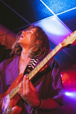 A person with long hair passionately plays an electric guitar under vibrant stage lighting. The musician wears a striped shirt, and the background features a colorful, blurred light effect, suggesting a lively performance setting.