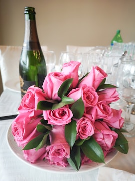 A bouquet of pink roses with lush green leaves is elegantly arranged on a white plate. In the background, a bottle of champagne and several wine glasses are visible, suggesting a celebratory or romantic setting.