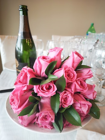 A bouquet of pink roses with lush green leaves is elegantly arranged on a white plate. In the background, a bottle of champagne and several wine glasses are visible, suggesting a celebratory or romantic setting.