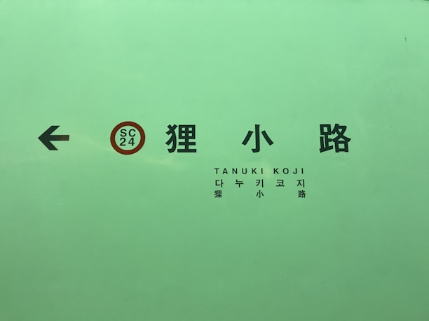 A green sign with multilingual text including Japanese, Korean, and Roman letters. There is an arrow pointing left and a circular SC 24 symbol.