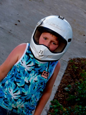 A child wearing a white motorcycle helmet looks directly at the camera. The child is dressed in a sleeveless shirt with a vibrant blue and white leaf pattern. The background consists of concrete pavement and some patches of greenery.