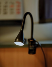 A focused image of a black, flexible-neck reading lamp emitting bright light, with a blurred background featuring a hint of a keyboard or control panel. The lamp is positioned over a wooden surface, creating a warm contrast.