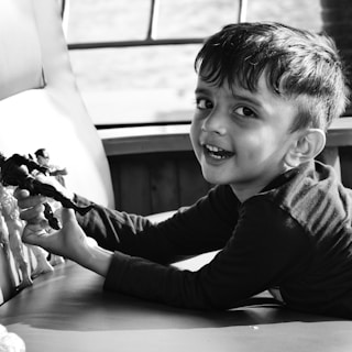 A young child with short hair is smiling while holding toy action figures. The child is sitting on a cushioned surface in front of a window. The setting is well-lit, creating a cheerful atmosphere.