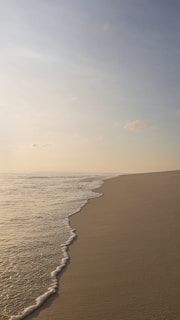 A serene sandy beach stretches into the distance, with gentle waves lapping at the shore under a calm, light blue sky. A soft, golden hue suggests a time around sunrise or sunset, casting a peaceful atmosphere.