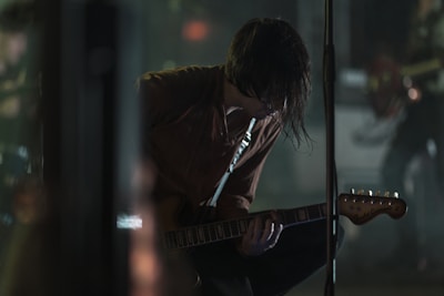 A musician crouches on stage, intensely focused, with long hair partially covering their face. They hold an electric guitar, suggesting a live music performance setting, with stage equipment visible in the background. The lighting is low and atmospheric, creating a moody, intimate ambiance.