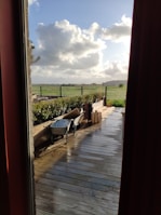 A wooden deck overlooks a green field with a partly cloudy sky. A wheelbarrow and some wooden planks are placed on the deck, indicating recent or ongoing work. The scene is viewed through a window frame and door curtains, adding a cozy indoor perspective.