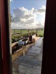 A wooden deck overlooks a green field with a partly cloudy sky. A wheelbarrow and some wooden planks are placed on the deck, indicating recent or ongoing work. The scene is viewed through a window frame and door curtains, adding a cozy indoor perspective.
