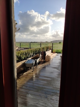 A wooden deck overlooks a green field with a partly cloudy sky. A wheelbarrow and some wooden planks are placed on the deck, indicating recent or ongoing work. The scene is viewed through a window frame and door curtains, adding a cozy indoor perspective.