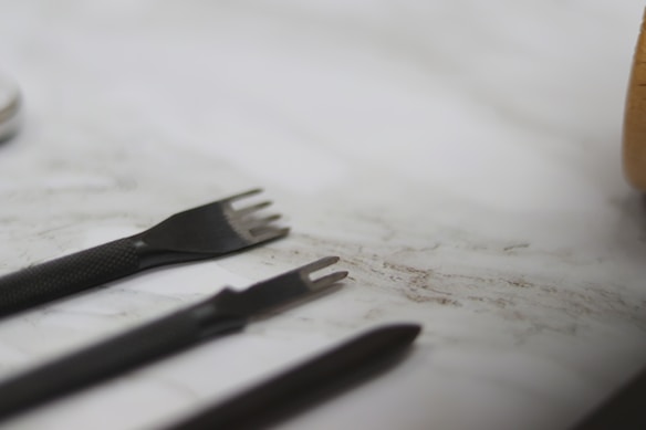 A set of black leather crafting tools, including two pronged punches and a pointed tool, are placed on a light marble surface. The focus is on the sharp ends of the tools, with the background slightly blurred.