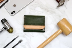 A green leather wallet with a brown accent placed on a marble surface is surrounded by various tools and an Apple smartphone. The tools include a wooden mallet, a metal hammer, and leather crafting tools.