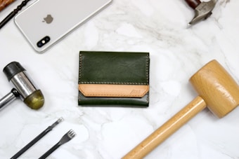 A green leather wallet with a brown accent placed on a marble surface is surrounded by various tools and an Apple smartphone. The tools include a wooden mallet, a metal hammer, and leather crafting tools.
