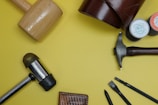 A variety of leatherworking tools are arranged on a yellow surface, including mallets, punches, needles, and thread spools. A piece of leather is partially shown in the upper right corner.
