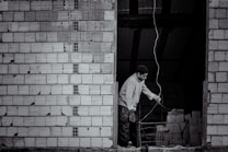A person stands near an unfinished brick wall, attentively working with wiring. The background shows a dark, unlit space with stacks of bricks.