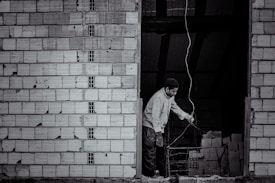 A person stands near an unfinished brick wall, attentively working with wiring. The background shows a dark, unlit space with stacks of bricks.