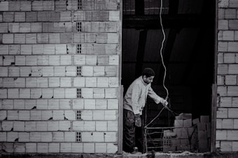 A person stands near an unfinished brick wall, attentively working with wiring. The background shows a dark, unlit space with stacks of bricks.