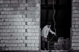 A person stands near an unfinished brick wall, attentively working with wiring. The background shows a dark, unlit space with stacks of bricks.
