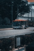 A public bus with the route number 195 is traveling on a tree-lined street in an urban area. The scene includes buildings with classical architecture in the background, and there are no other vehicles or pedestrians visible on the road.