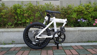 A compact, white folding bicycle stands on a tiled path with a background of neatly trimmed green shrubs and some flowering plants. The bicycle's black wheels and handlebar complement the white frame, giving it a sleek look. The setting conveys a tidy outdoor environment, possibly a park or garden.