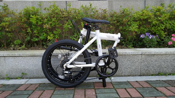 A compact, white folding bicycle stands on a tiled path with a background of neatly trimmed green shrubs and some flowering plants. The bicycle's black wheels and handlebar complement the white frame, giving it a sleek look. The setting conveys a tidy outdoor environment, possibly a park or garden.