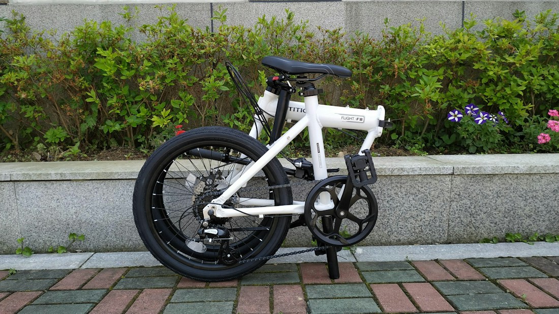 A compact, white folding bicycle stands on a tiled path with a background of neatly trimmed green shrubs and some flowering plants. The bicycle's black wheels and handlebar complement the white frame, giving it a sleek look. The setting conveys a tidy outdoor environment, possibly a park or garden.