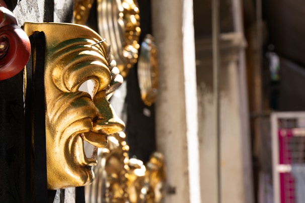 A close-up view of a shiny gold theatrical comedy mask rested on a textured black surface. The mask features exaggerated facial expressions, highlighting a joyful or humorous theme. Additional golden masks are blurred in the background, suggesting a display or collection.