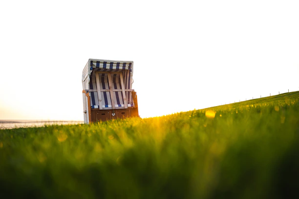 A beach chair with a striped pattern is placed on a grassy field, with sunlight illuminating the scene from the background. The focus is on the chair, while the foreground and background are softly blurred, creating a serene and peaceful atmosphere.