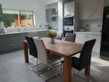 A modern kitchen features a wooden dining table surrounded by six chairs in various colors. The kitchen is equipped with sleek cabinets, a built-in oven, and a gas stove. Natural light streams in through a window adorned with blinds, illuminating potted plants on the windowsill. A fruit bowl and a vase with yellow flowers serve as decorative elements.