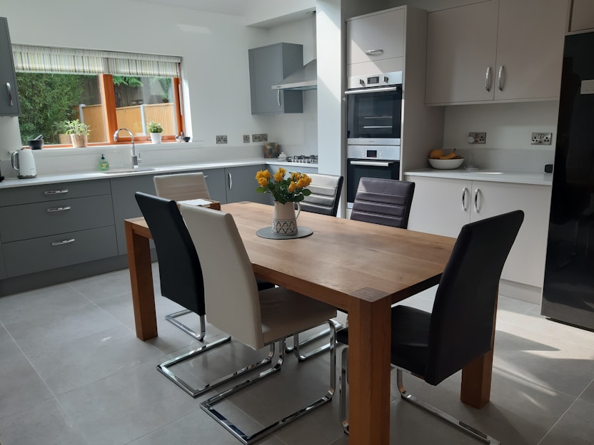 A modern kitchen features a wooden dining table surrounded by six chairs in various colors. The kitchen is equipped with sleek cabinets, a built-in oven, and a gas stove. Natural light streams in through a window adorned with blinds, illuminating potted plants on the windowsill. A fruit bowl and a vase with yellow flowers serve as decorative elements.