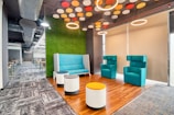 A modern office lounge area features contemporary seating, including a blue couch and two teal armchairs. The space is enriched by circular hanging lights and a unique ceiling design with colorful circular decorations. A green carpeted wall adds an element of nature, complemented by a brown wooden floor. Adjacent to this space is a hallway lined with workstations, under industrial-style air ducts.