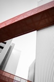 A modern architectural structure featuring tall buildings and a prominent red metal bridge or walkway connecting them. The lines are clean with a focus on industrial design elements, incorporating vertical corrugated panels in a muted gray tone.