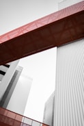 A modern architectural structure featuring tall buildings and a prominent red metal bridge or walkway connecting them. The lines are clean with a focus on industrial design elements, incorporating vertical corrugated panels in a muted gray tone.